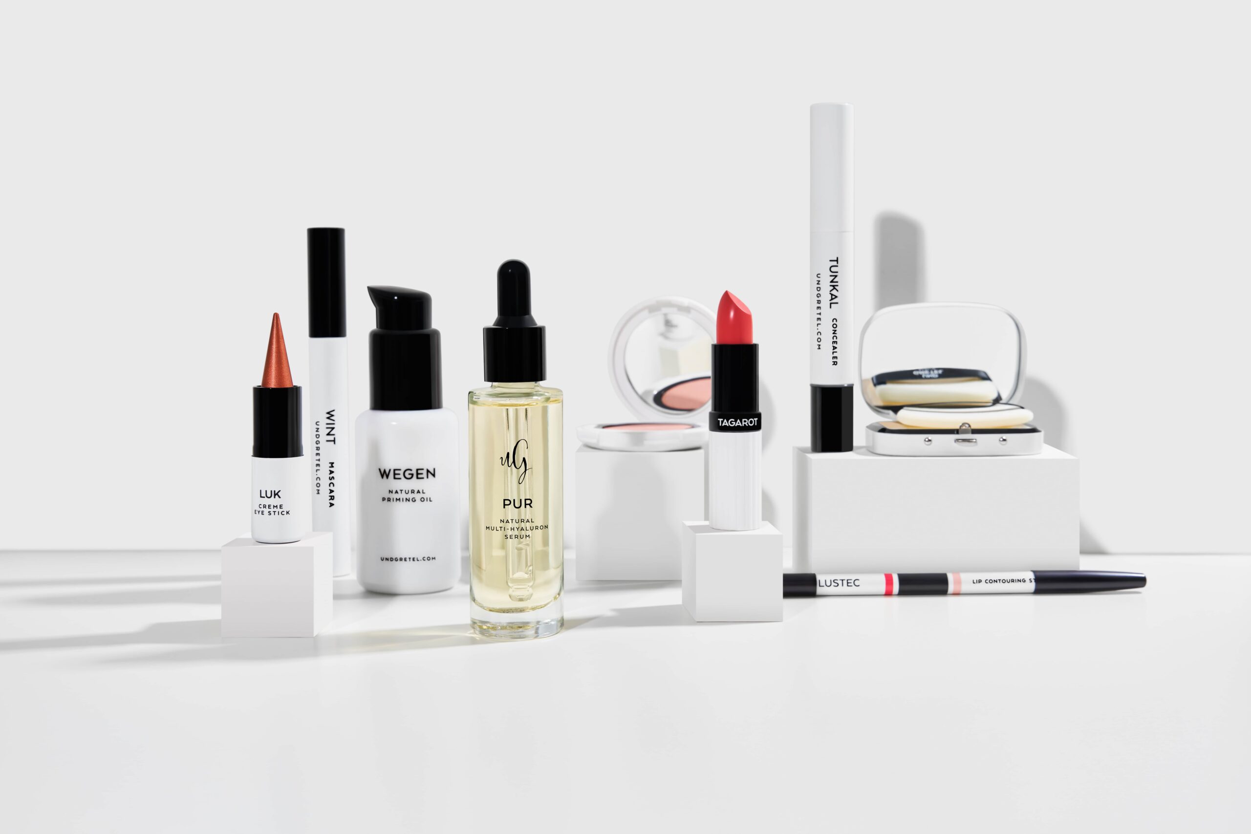 Arrangement von neun UND GRETEL Naturkosmetik-Produkten in minimalistischen Flaschen und Verpackungen auf weißen Podesten, inklusive Serum, Mascara, Lippenstift und Concealer.